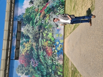Rowan standing in front of a large mural of a woodland and a pond, painted on a large building