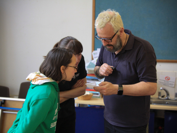 Two people leaning over to examine a specimen tray being held by a third person in an indoor setting