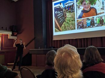 Vicky stood in front of a group of people talking with a presentation projected onto a drop down screen
