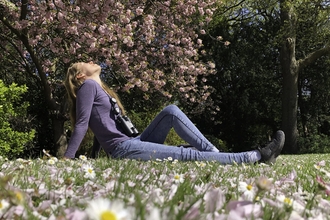 A beautiful young woman sits on the grass in a park, with daisies in the foreground and blossoming trees behind. She has her head thrown back and her eyes closed, enjoying the peace of nature