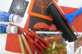Linocutting tools, paint, a picture of a mushroom, and some printed flowers and mushrooms overlapping each other on a table