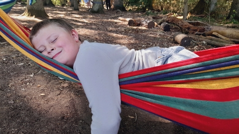 A young boy laying across a rainbow coloured hammock with a big smile