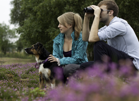 A young man and woman sit amongst flowering heather, with a dog on a lead sat beside them. The woman has her hands on the dog, whilst the man looks through a pair of binoculars.